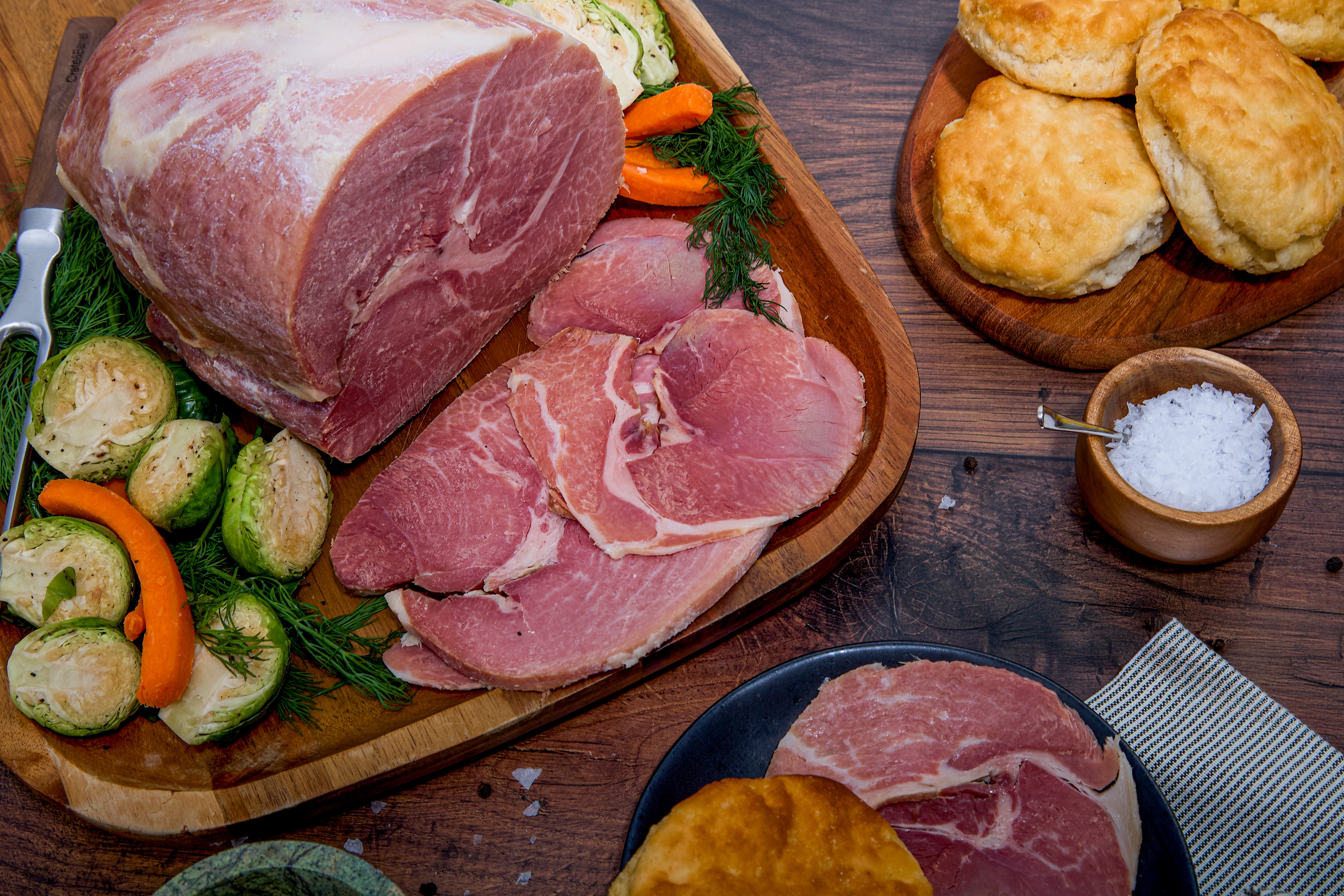 Wooden cutting board with sliced Turner Ham, vegetables, and biscuits on a wooden table.