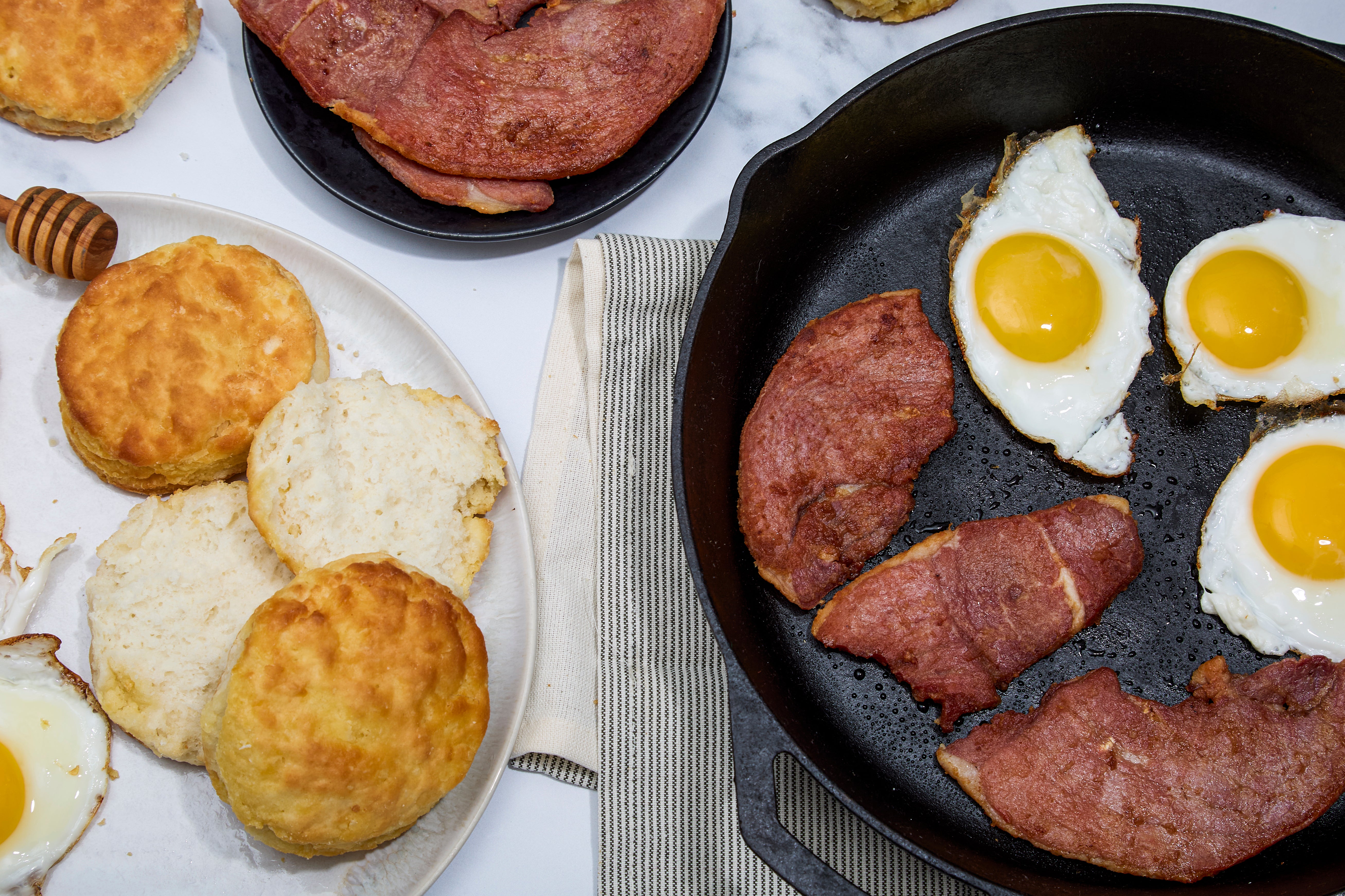 Breakfast setting with biscuits on a white plate and slices of Turner Ham, and eggs in a skillet.