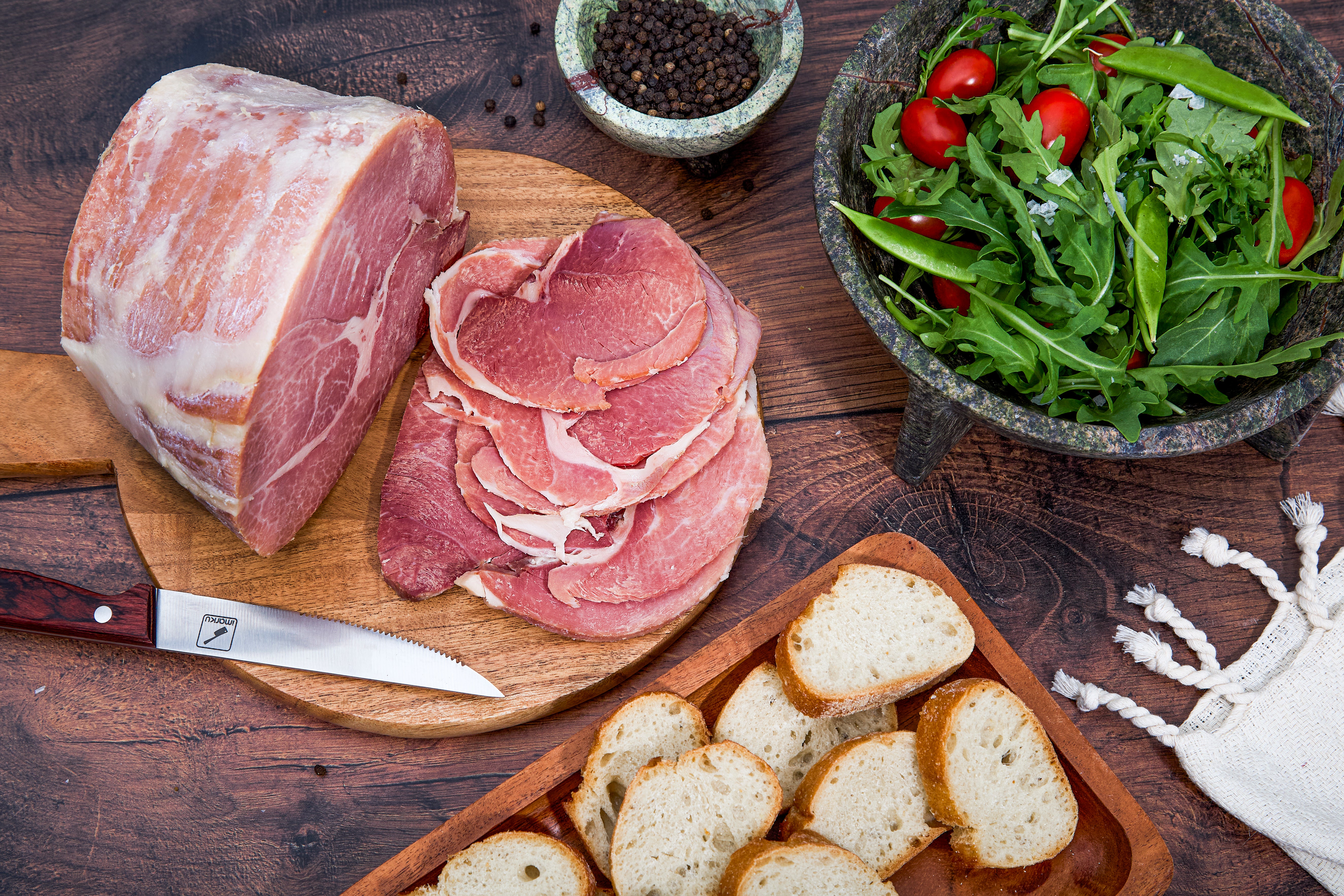 Close up of a dinner table with sliced Turner Ham on a cutting board surrounded by slices of bread, peppercorns, and a bowl of salad.