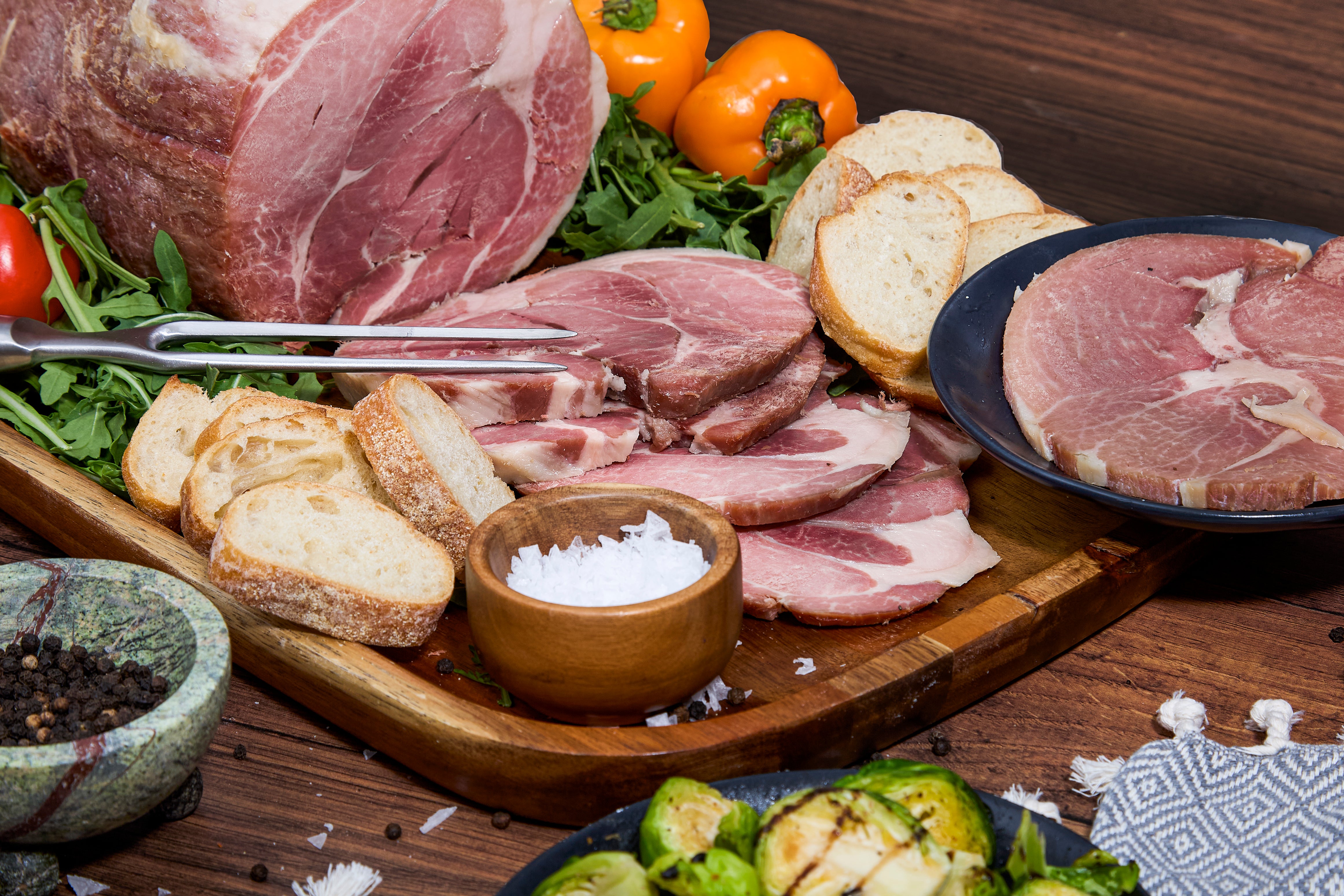 Platter of sliced cured Turner Ham with bread, vegetables, and a bowl of salt on a wooden surface.