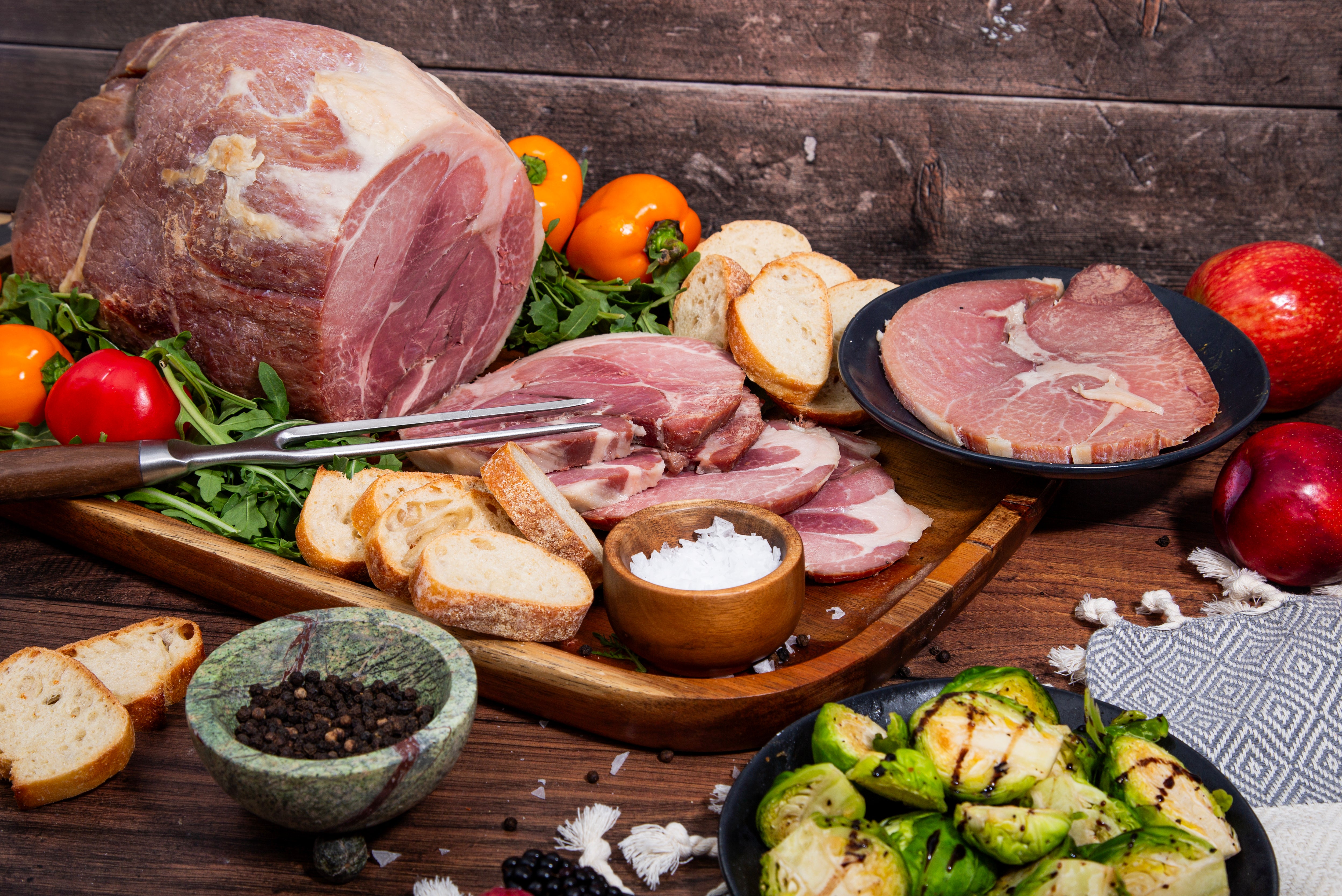 A whole cooked Turner Ham partially sliced on a serving board surrounded by assorted fruits and vegetables.